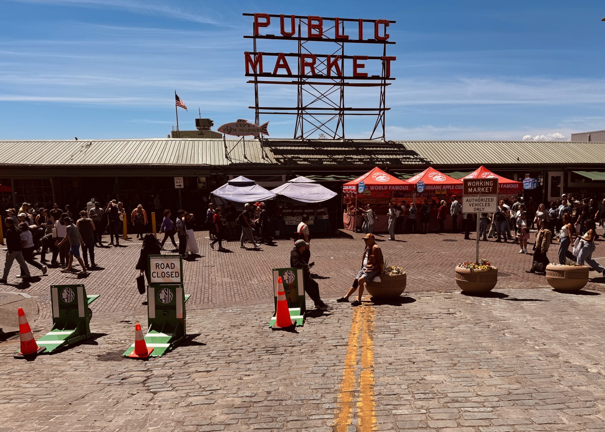 A wide cobblestone plaza at Seattle’s Pike Place Market is busy with pedestrians, vendor tents. Road barriers and cones restrict vehicle access. The iconic red Public Market sign towers above the market building, and bright red cider tents line one side of the scene under a clear blue sky.