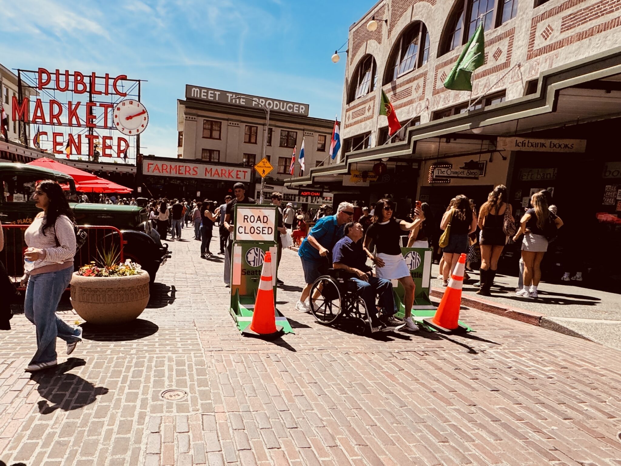 A busy scene at Seattle’s Pike Place Market shows pedestrians moving along a cobblestone street closed to vehicles with cones and a barriers. In the center, a small group pauses near a barrier, with one woman appearing to take a selfie beside a man in a wheelchair, while the iconic Public Market Center sign and market buildings rise in the background under a bright blue sky.