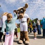 A Seattle Mariners moose mascot high-fives a young girl while families gather around on a sunny outdoor walkway. Children and adults watch the playful interaction during what appears to be a community event.