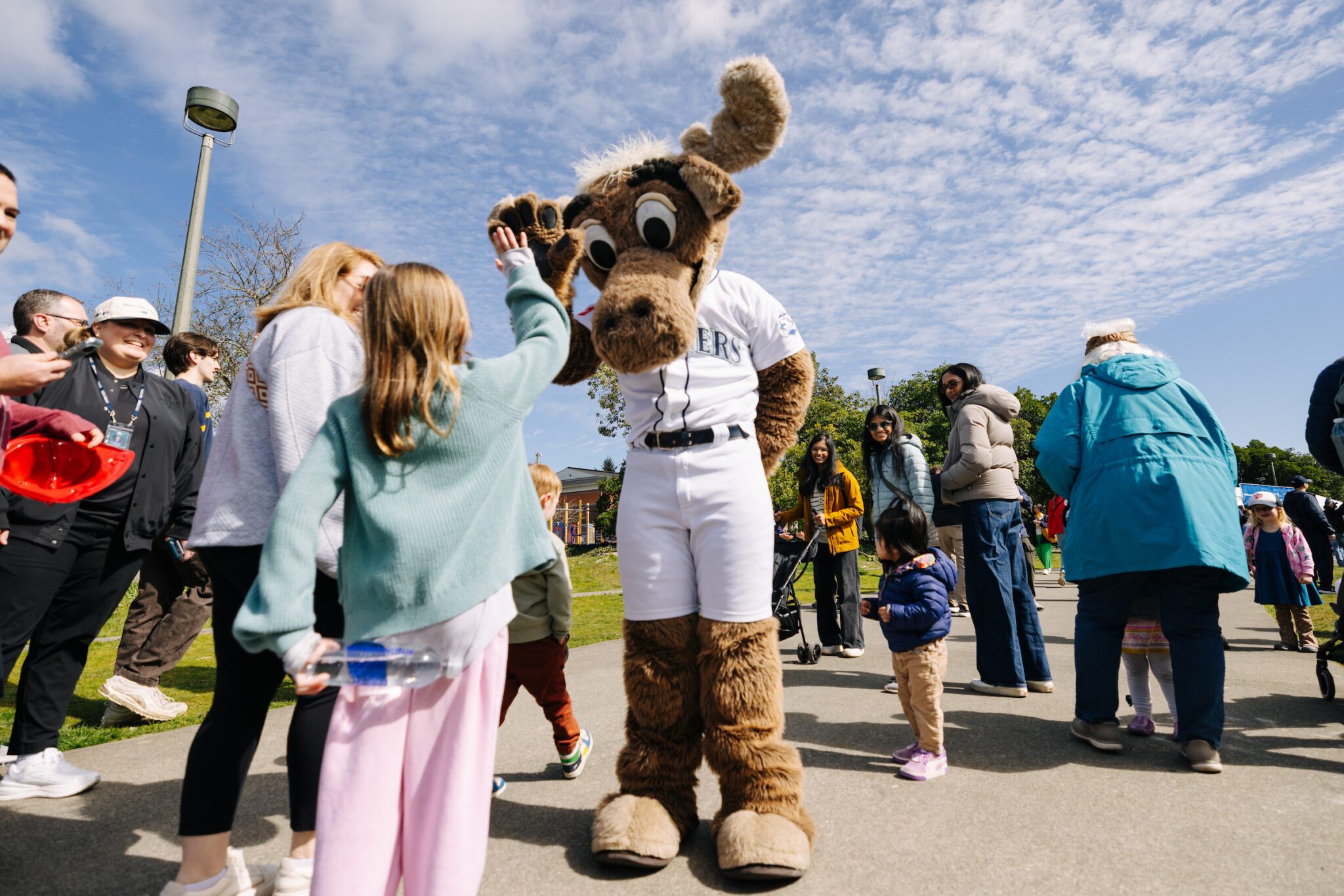 A Seattle Mariners moose mascot high-fives a young girl while families gather around on a sunny outdoor walkway. Children and adults watch the playful interaction during what appears to be a community event.