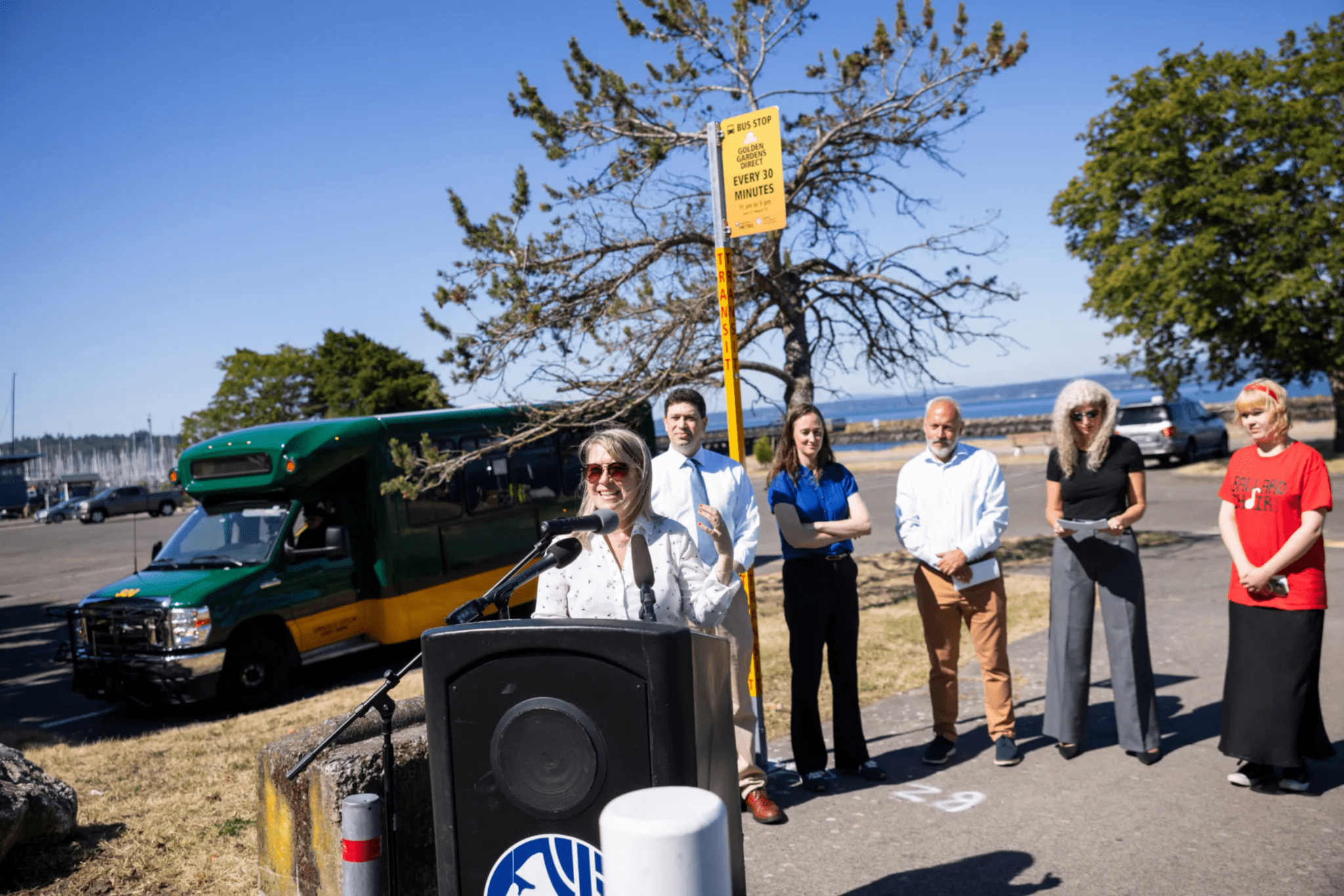 Woman speaks at a podium outdoors beside a green-and-yellow shuttle bus while five people stand behind her near a sign reading, “Bus Stop, Golden Gardens Direct, Every 30 Minutes, 11 am to 9 pm,” with water and a clear blue sky in the background.