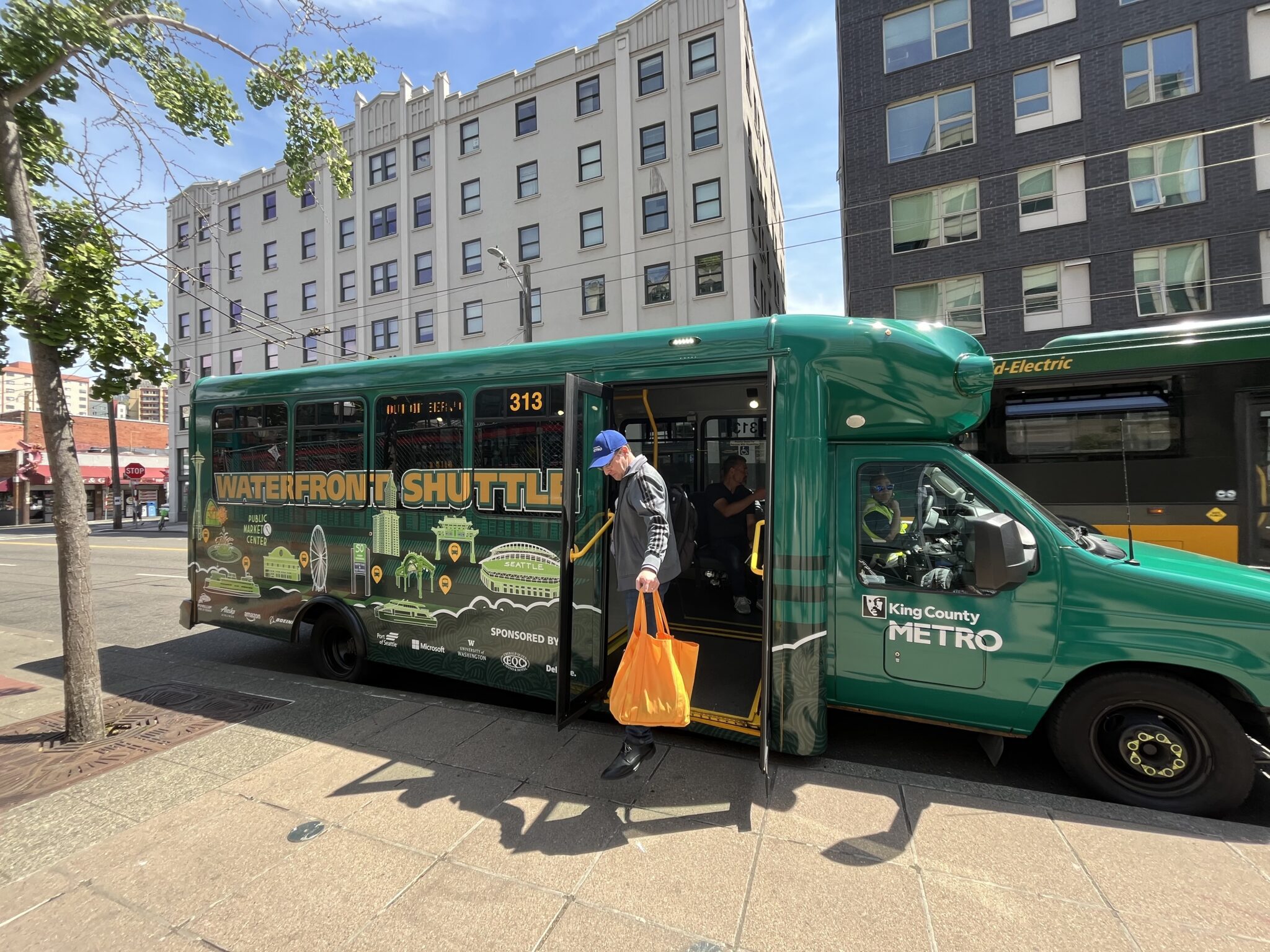 A green Seattle Waterfront Shuttle is stopped at a city curb as a man carrying a bright orange bag steps off through the open door. Another bus is parked behind it, and tall downtown buildings, a stop sign, and overhead wires frame the sunny street scene.