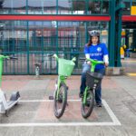 A woman stands with a green shared e-bike in a marked parking area. She wears a helmet and a blue City of Seattle shirt. A second green bike and a white-and-green scooter are beside her. A transit station is in the background.