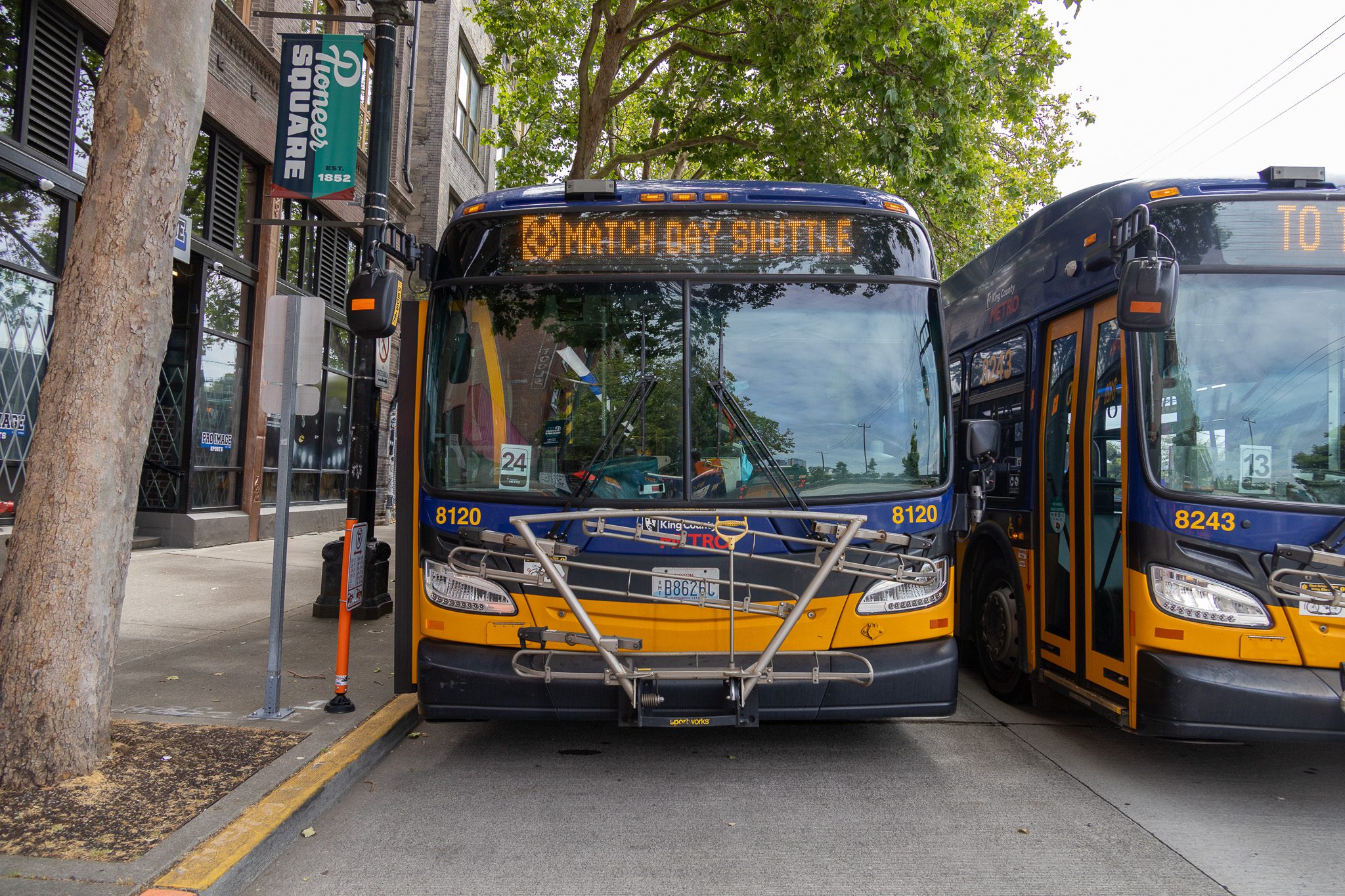 Two King County Metro buses are parked along a curb in Seattle’s Pioneer Square neighborhood beneath leafy trees. The bus in front displays “Match Day Shuttle,” and a green street banner reads “Pioneer Square Est. 1852.”