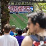A crowd gathers outdoors in a tree-lined plaza to watch a live soccer match on a giant video screen. A mossy tree trunk and nearby spectators frame the scene, emphasizing the shared public watch-party atmosphere.