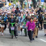A crowded city parade moves down the street toward the camera. In front, two people ride bright green electric seated scooters beside event staff in safety vests. Behind them, two police officers are on horseback, one is holding an American flag.
