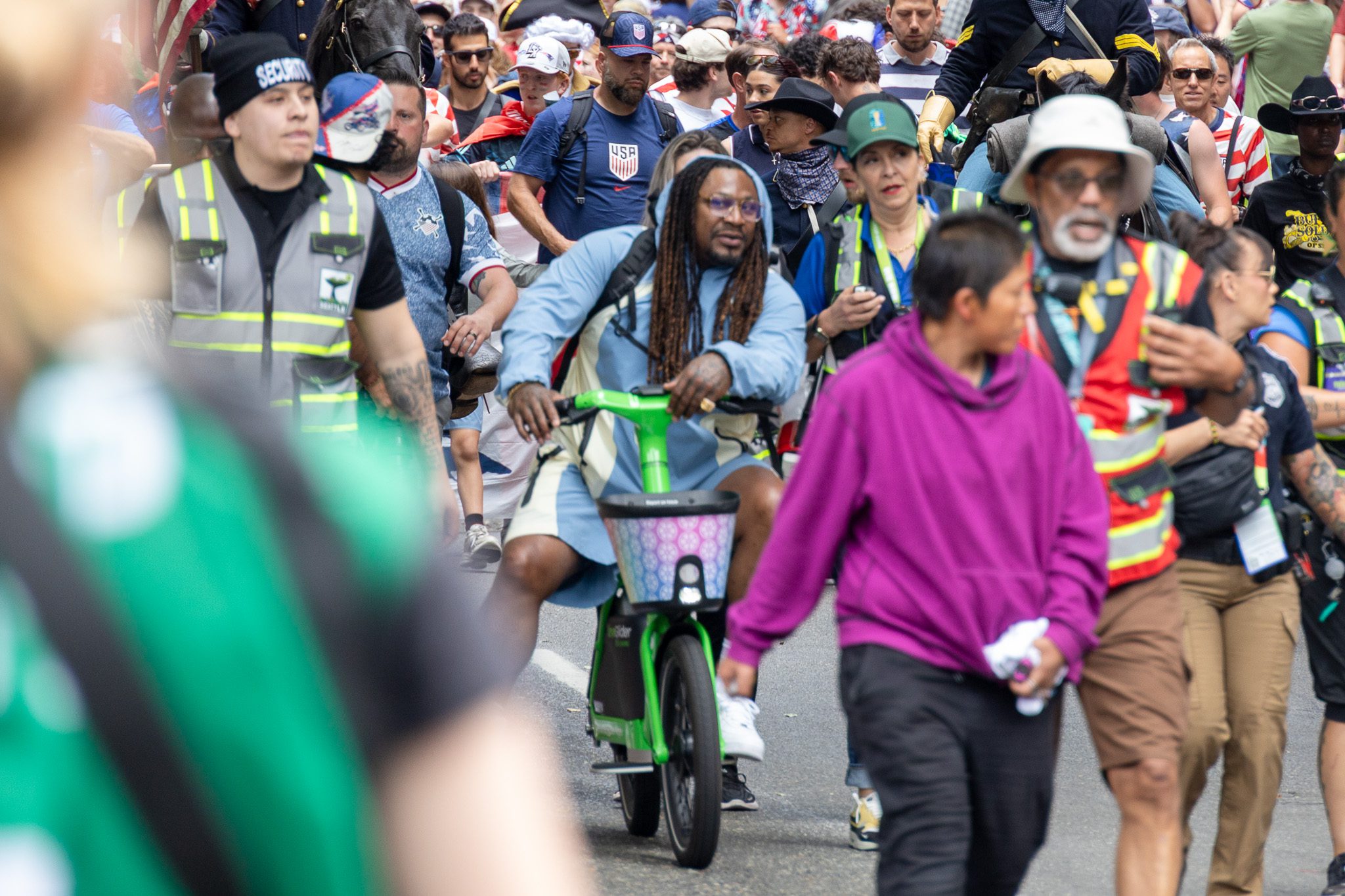 A crowded parade moves down a city street as a man rides a bright green scooter through the center of the scene, surrounded by event staff, pedestrians, and a large crowd. In the foreground, a person in a purple hoodie walks past, while security staff and a horse in the background suggest an organized public celebration.