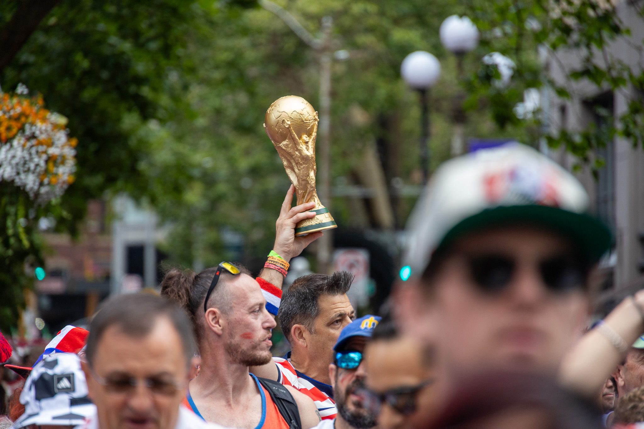 A golden World Cup trophy replica is held above a tightly packed crowd of soccer fans celebrating outdoors, with people in team colors and hats gathered beneath trees on a city street.