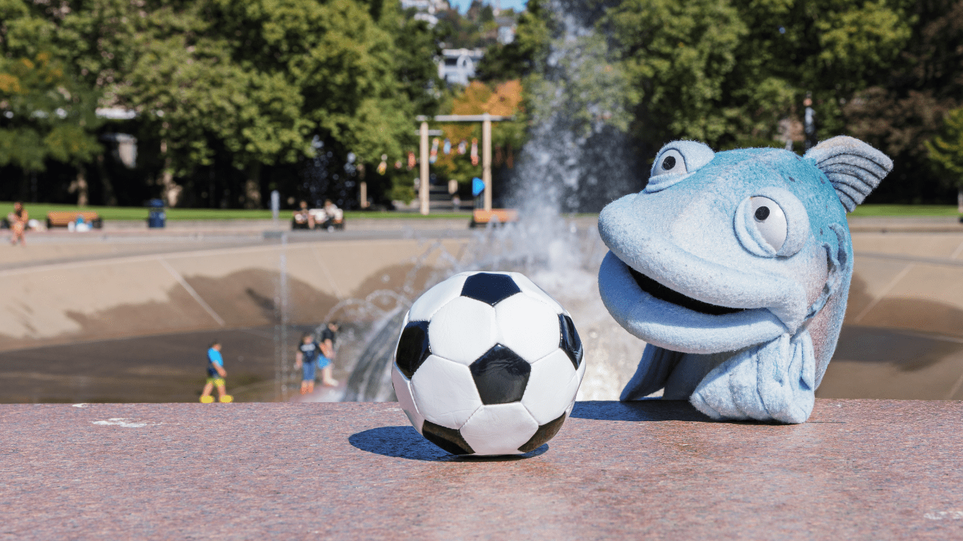 A blue fish puppet and a soccer ball sit on a stone ledge in front of a fountain at a sunny park.
