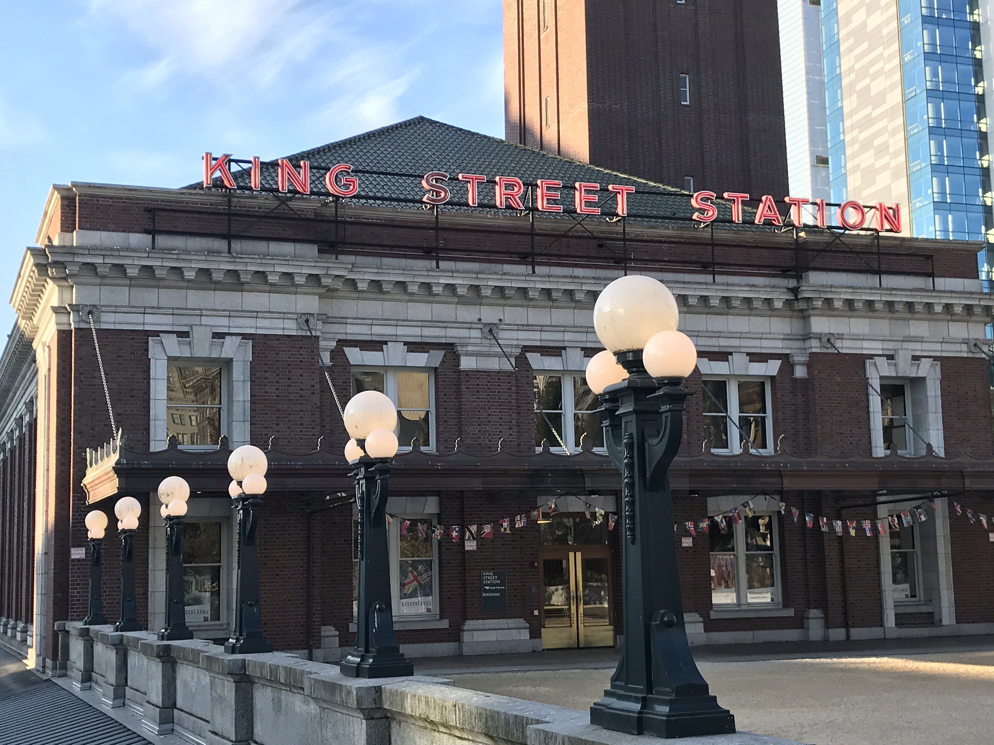 A historic brick train station stands in daylight, with a red rooftop sign that reads King Street Station. A row of black vintage-style lamp posts topped with white glass globes lines the foreground walkway. Behind the station, a dark brick tower and a modern glass building rise into a clear blue sky.