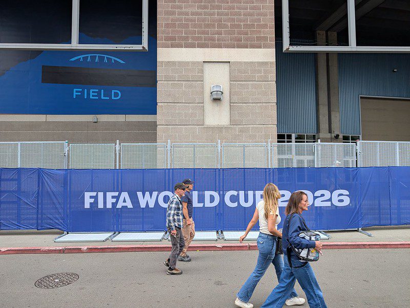 Exterior of a stadium with a blue fence banner reading “FIFA WORLD CUP 2026” as four people walk past on the sidewalk