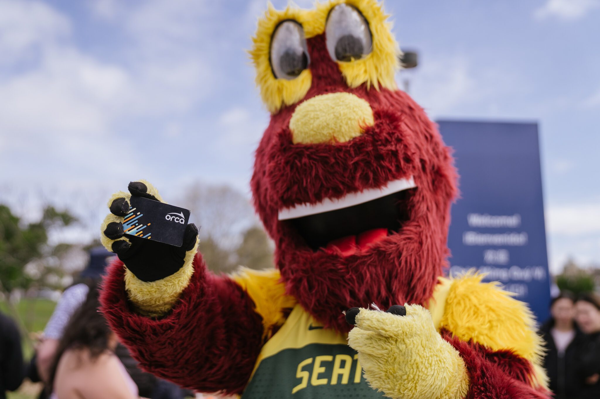 A purple mascot in a Seattle Storm jersey smiles at the camera while holding up a transit card that says 