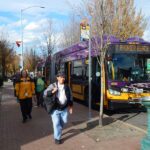 Purple and yellow Route 7 bus to Rainier Beach stopped with doors open at a city bus stop as four people walk along the sidewalk.