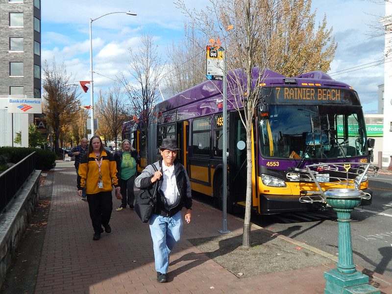 Purple and yellow Route 7 bus to Rainier Beach stopped with doors open at a city bus stop as four people walk along the sidewalk.