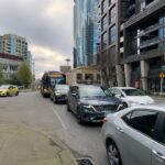 Street-level view of a busy street on an overcast day. A line of vehicles sits in traffic, including a blue and yellow King County Metro bus on Route 8 to Mt. Baker Transit Center. Modern glass high-rise buildings rise in the background, and the Seattle Space Needle appears in the distant center.