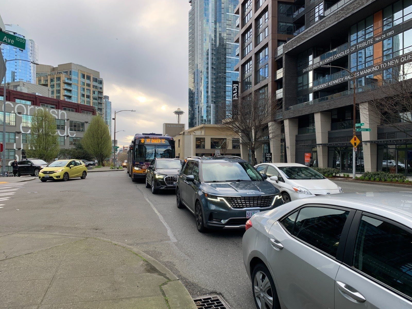 Street-level view of a busy street on an overcast day. A line of vehicles sits in traffic, including a blue and yellow King County Metro bus on Route 8 to Mt. Baker Transit Center. Modern glass high-rise buildings rise in the background, and the Seattle Space Needle appears in the distant center.