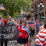 A crowd of US soccer supporters in red, white and blue march down S Washington St in Seattle. A smiling woman holds an American flag and wears a Seattle scarf. A black banner reads "DON'T GIVE UP THE SHIP."