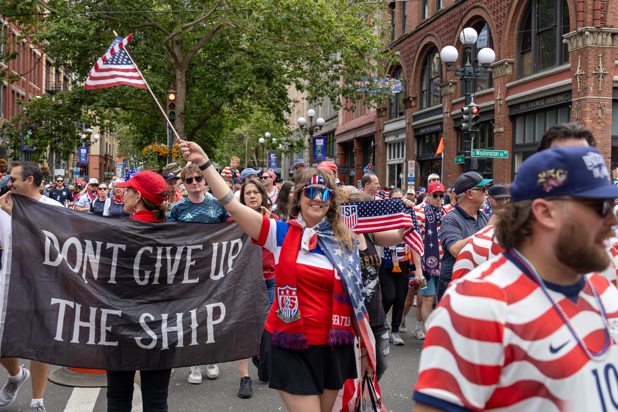 A crowd of US soccer supporters in red, white and blue march down S Washington St in Seattle. A smiling woman holds an American flag and wears a Seattle scarf. A black banner reads 