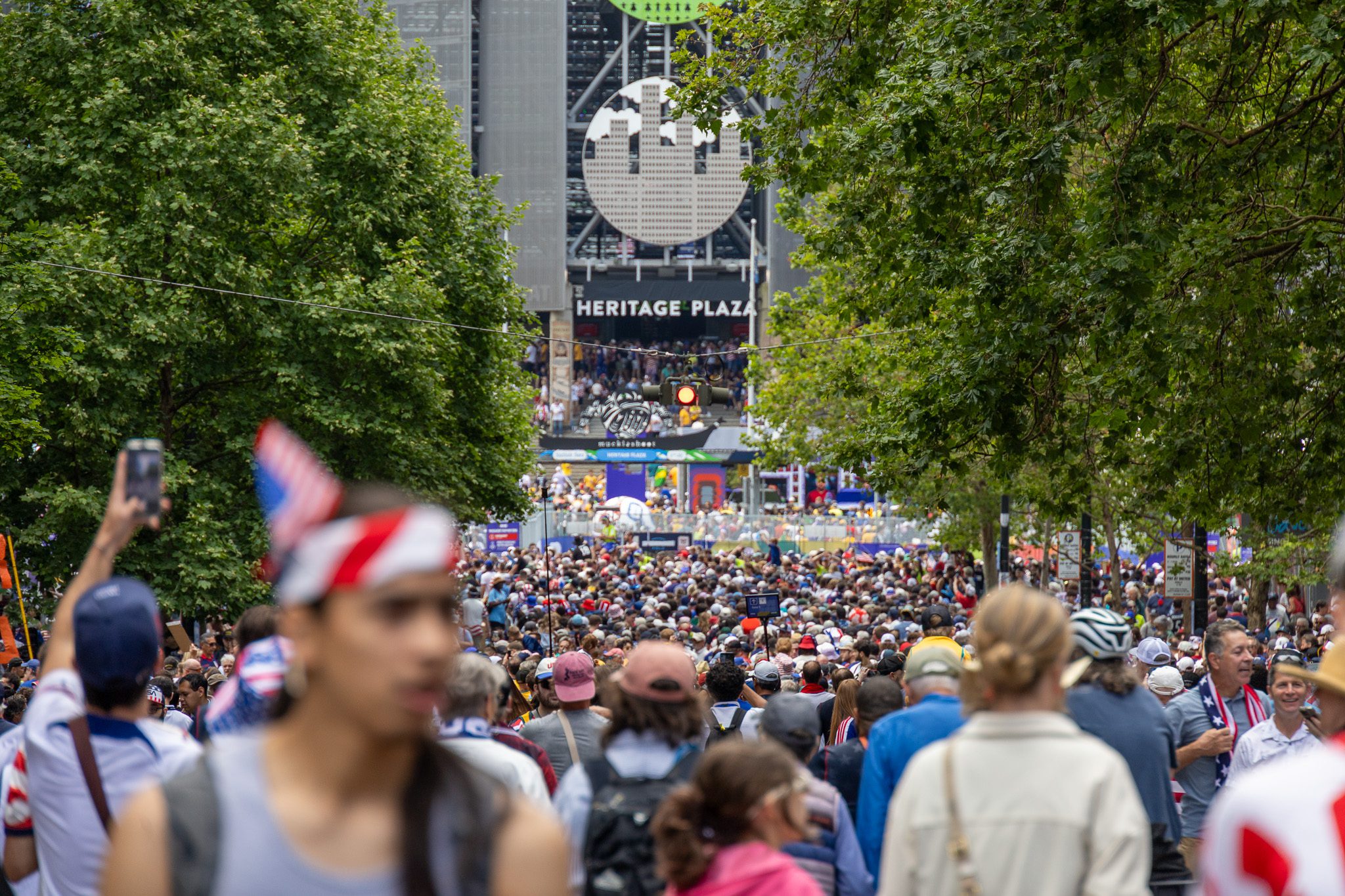 A large crowd of people in red, white, and blue clothing walks down a tree-lined path toward the entrance of a stadium.