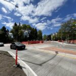 A road intersection or roundabout under construction on Roosevelt Way, with a black car driving through curved concrete lanes, orange barriers and cones, a worker in a safety vest, trees, and a bright blue sky with scattered clouds.