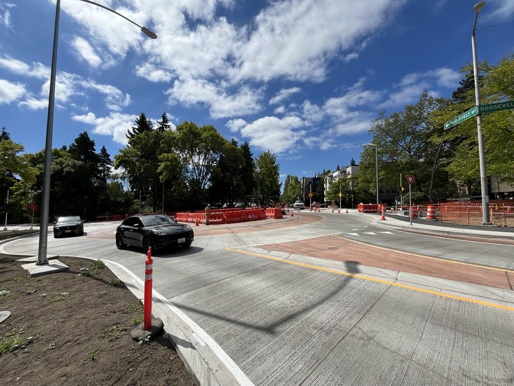 A road intersection or roundabout under construction on Roosevelt Way, with a black car driving through curved concrete lanes, orange barriers and cones, a worker in a safety vest, trees, and a bright blue sky with scattered clouds.
