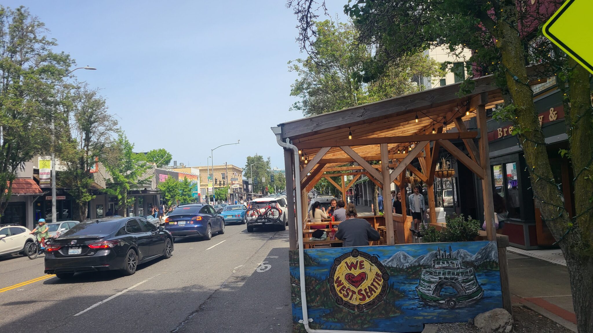 Sunny neighborhood business district with cars, a cyclist, and pedestrians along a main street beside a wooden outdoor dining patio painted with a West Seattle mural.