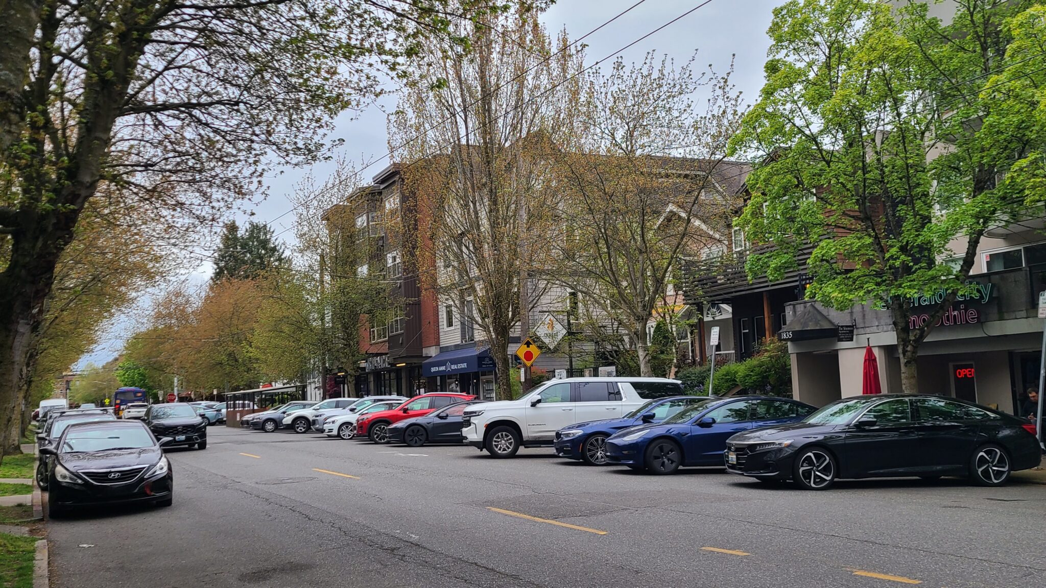 Tree-lined neighborhood street with angled parked cars beside storefronts and apartments, including Queen Anne Real Estate and Emerald City Smoothie, on an overcast day.