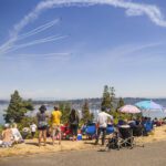 A crowd of people sit and stand on a dry, grassy hillside overlooking a wide lake on a sunny summer day. They are watching several jets fly in formation across a bright blue sky, leaving long white streaks of smoke behind them.
