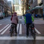 Two cyclists—a woman in a pink quilted jacket and dark-red helmet, and a man in a dark blue jacket with neon-green sleeves—wait at a crosswalk, seen from behind. Pedestrians walk the sidewalk to their right. A street sign reads 3rd Ave, near Pine St.