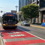 A bus travels in a red bus lane with cars, trees, people, and buildings in the background. The Space Needle is also in the background.