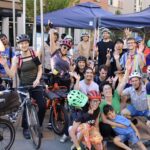 A large group of people celebrate at a bike event. Many of the people are wearing bike helmets and the event takes place outdoors.