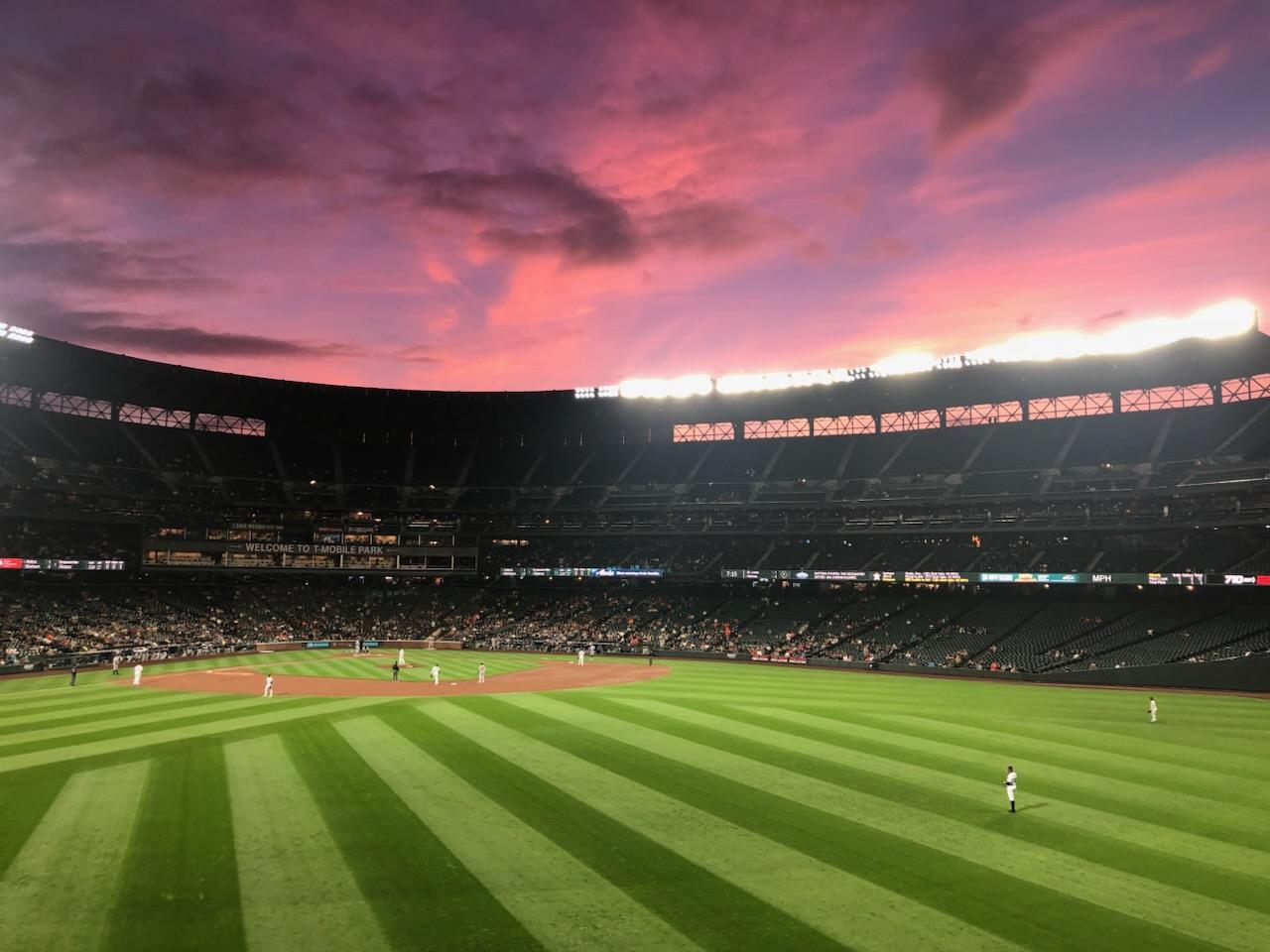A wide angle view of a baseball stadium in the evening. Green grass is shown below along with pink clouds in the sky, and crowds sitting in the stadium.