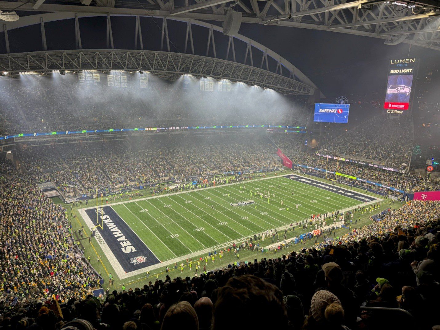 A full football stadium on a dark evening night with many fans cheering on a team and green turf in the center of the image.