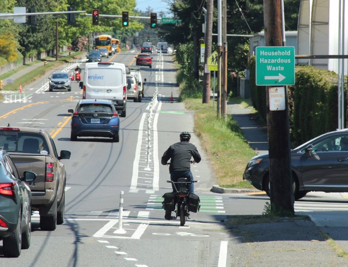 A person bikes in a protected bike lane with cars traveling to the left and right. School buses are in the background.
