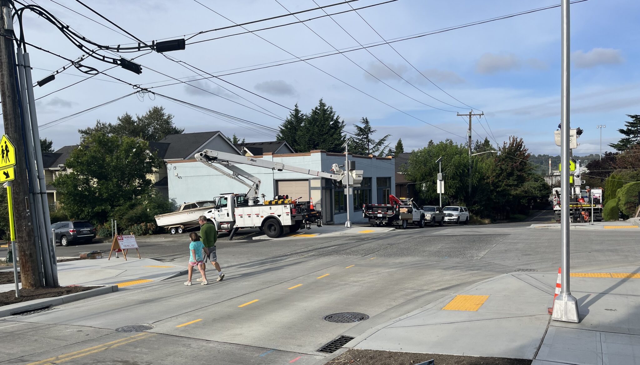 Two people cross the street. A work truck and other parked vehicles are in the image, as well as a light blue building and blue skies above.