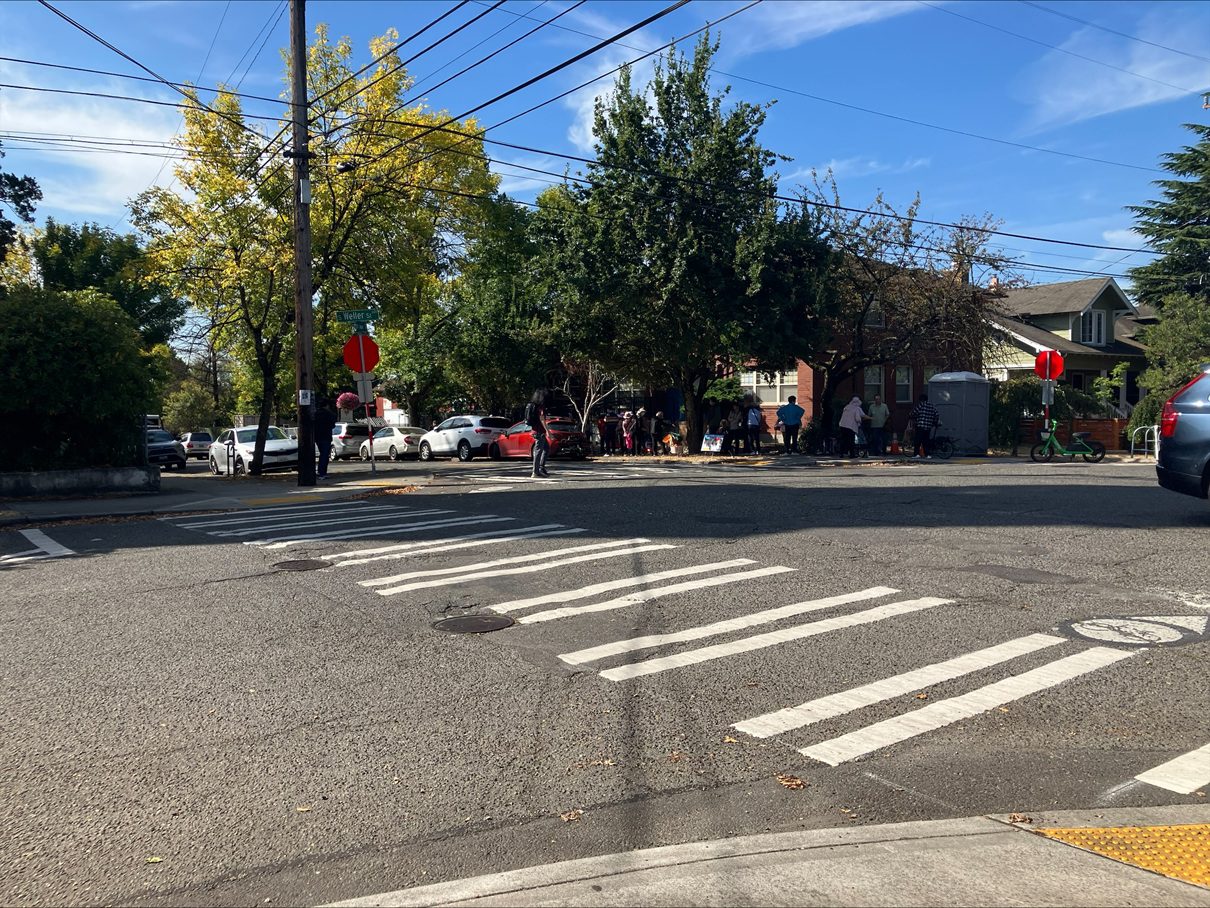 Marked crosswalks along a street with buildings and trees and people in the background.