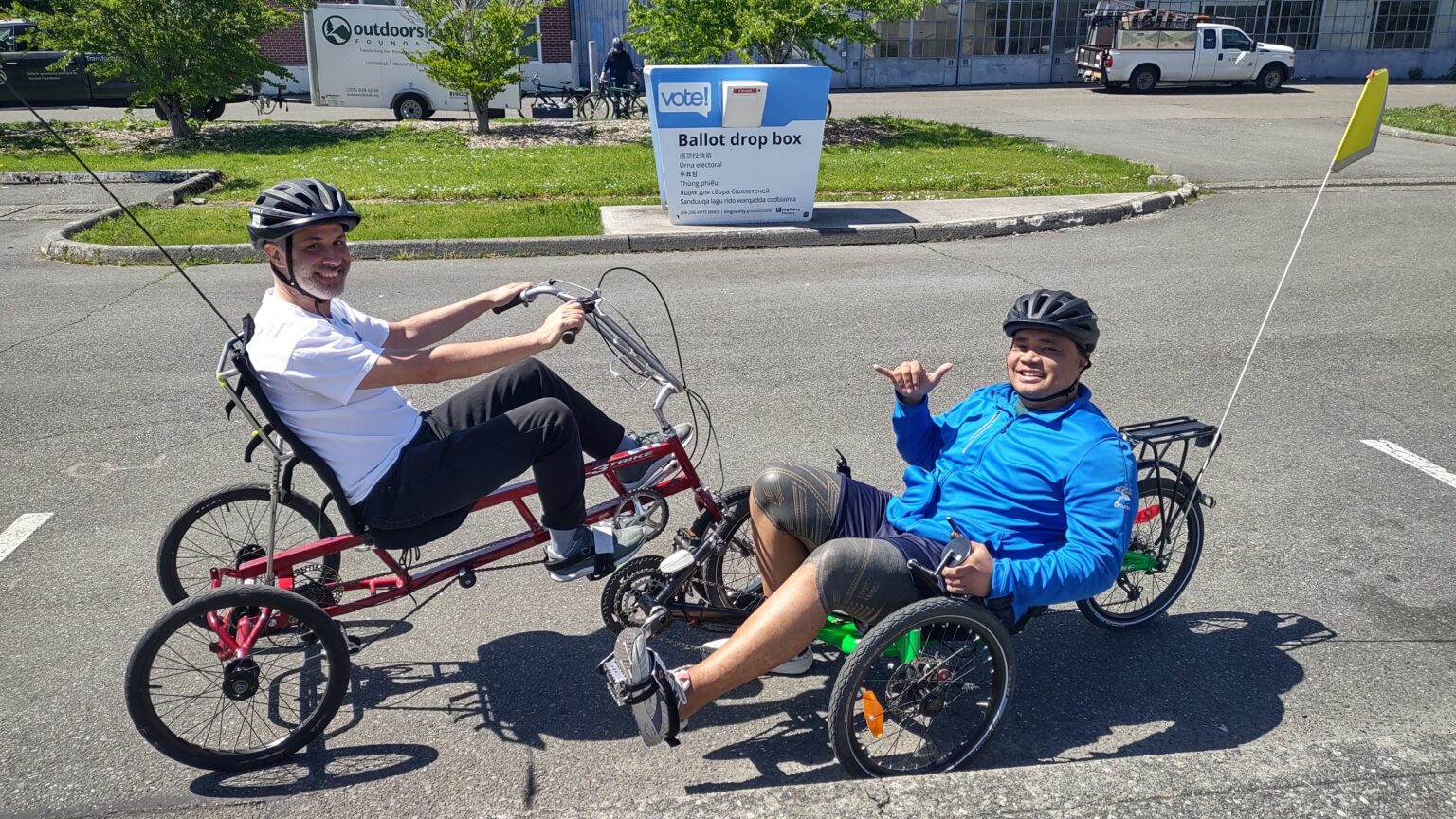 Two people smile while riding adaptive cycles in a parking lot area.