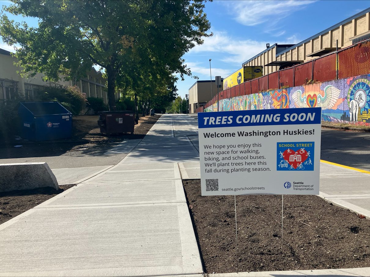A photo of expanded sidewalks with a large building and tree and dumpsters in the background. An informational yard sign says that trees are coming soon and welcomes Washington Huskies.