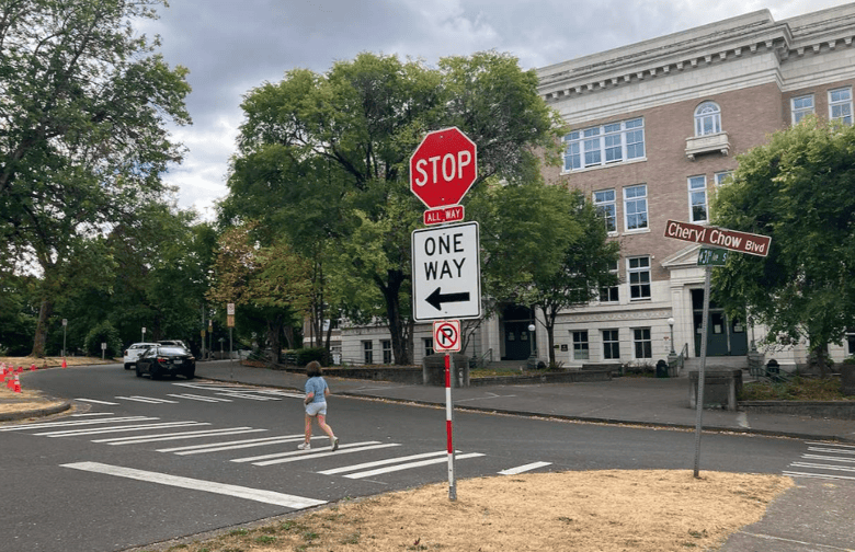 A young person crosses the street in front of a large school building at a marked crosswalk. A red stop sign, a one-way sign, and a street sign reading Cheryl Chow Blvd are in the image, as well as trees and parked cars.