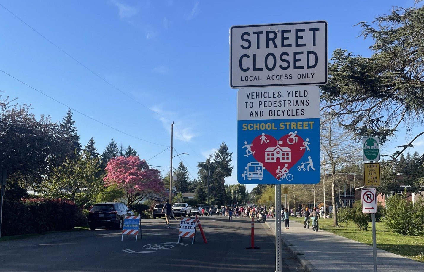 A School Street sign notes that the street is closed and local access only. A sign below says that vehicles yield to pedestrians and bicycles. A large group of people are in the background along with trees and parked cars.