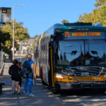 A King County Metro bus with a sign reading "75 Northgate Station" arrives at a bus stop on a bright, sunny day. Three people wait on a concrete sidewalk. Green and yellowing autumn trees fill the background under a clear blue sky.