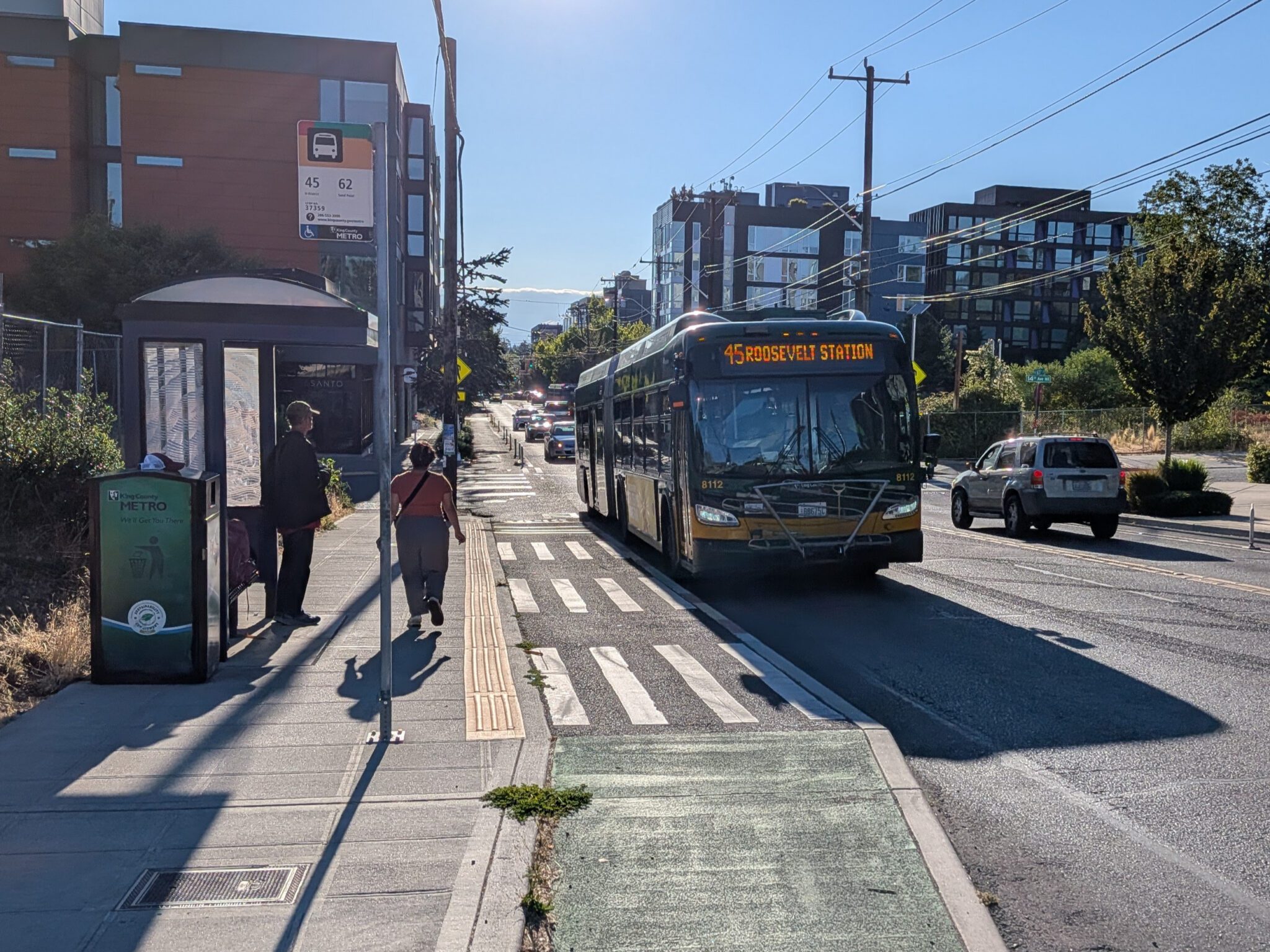 A bus with a sign reading “45 Roosevelt Station” pulls up to a bus stop on a bright, sunny day. One person waits under a covered bus shelter while another walks away along the sidewalk. A raised bike lane runs between the sidewalk and the bus. In the background are passing cars, modern apartment buildings, trees, and overhead utility wires against a clear blue sky.