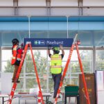 Two construction workers in hard hats and safety vests install a dark blue transit sign that says "1" in a green circle, "2" in a blue circle, "44" and "Pinehurst" while standing on red ladders at an outdoor station platform under construction.