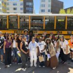 A large group of people stand in front of a yellow school bus on a sunny day.