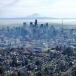 An aerial view of a city, with large and small buildings in the background, as well as a mountain and partly cloudy skies above.