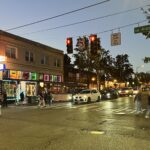 Several people cross the street in the early evening. Large buildings, cars, and traffic signals are in the background.