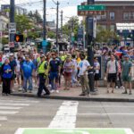 A large group of people at a sidewalk prepare to cross the street.