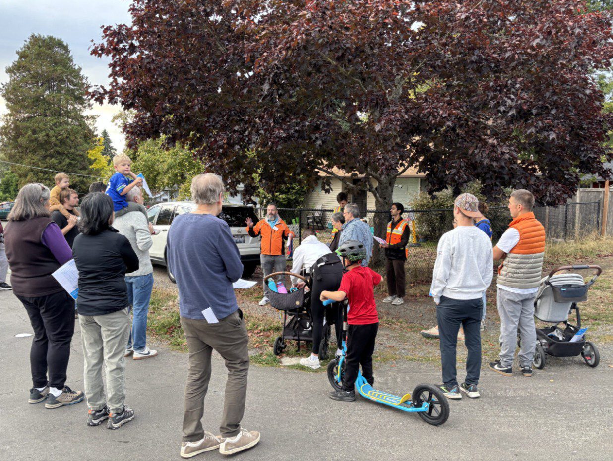 A large group of people walk along a street while discussing information. A large tree and houses are in the background.