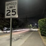 A photo of a 25 mph speed limit sign at night. Parked cars are in the background as well as a large hedge and the street in the center of the image.