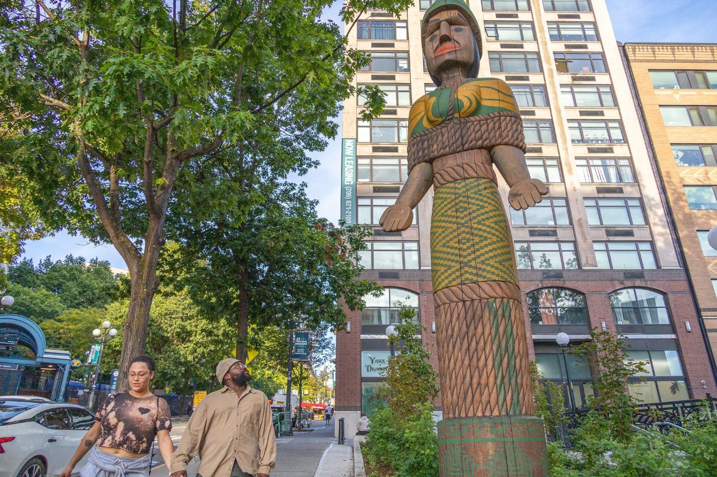 Two people look up at a large statue in the city. Tall buildings and trees are in the background.